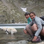 Two women hiking kneeling down next to golden retriever dog in a lake in the mountains 