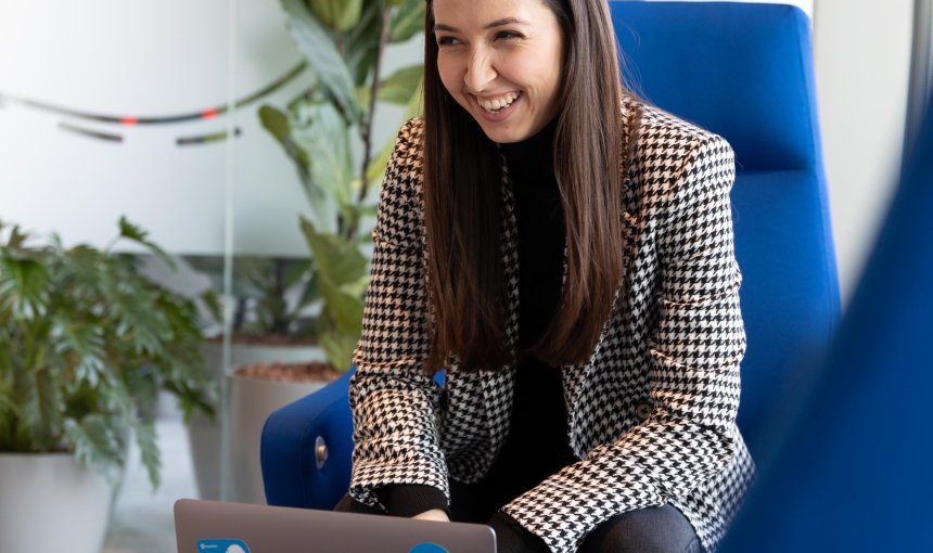 woman with brown hair sitting in a blue chair with laptop laughing