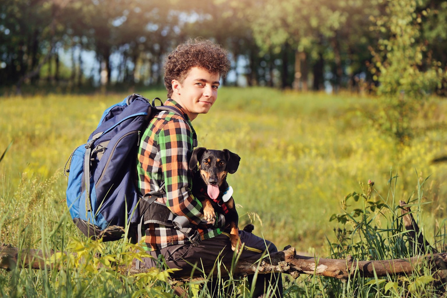 Hundebesitzer sitzt mit Rucksack und Hund auf dem Schoß in der Wiese auf einem Ast