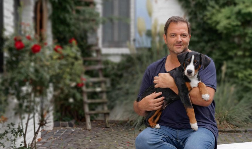 Martin Rütter sitzt vor einem Haus mit einem Welpen auf dem Schoß