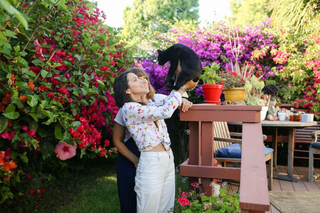 Two women play with a cat outdoors in a garden full of potted plants.