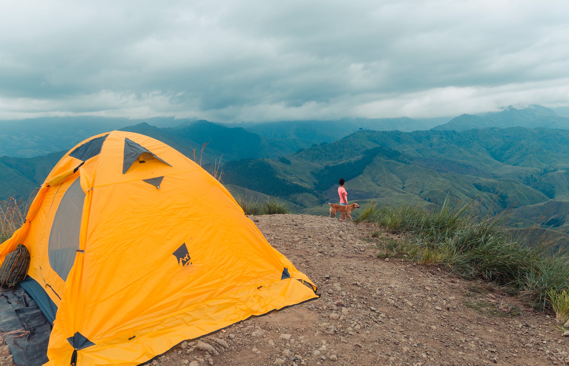 A woman walking with her dog in the mountains with a yellow tent set up behind them