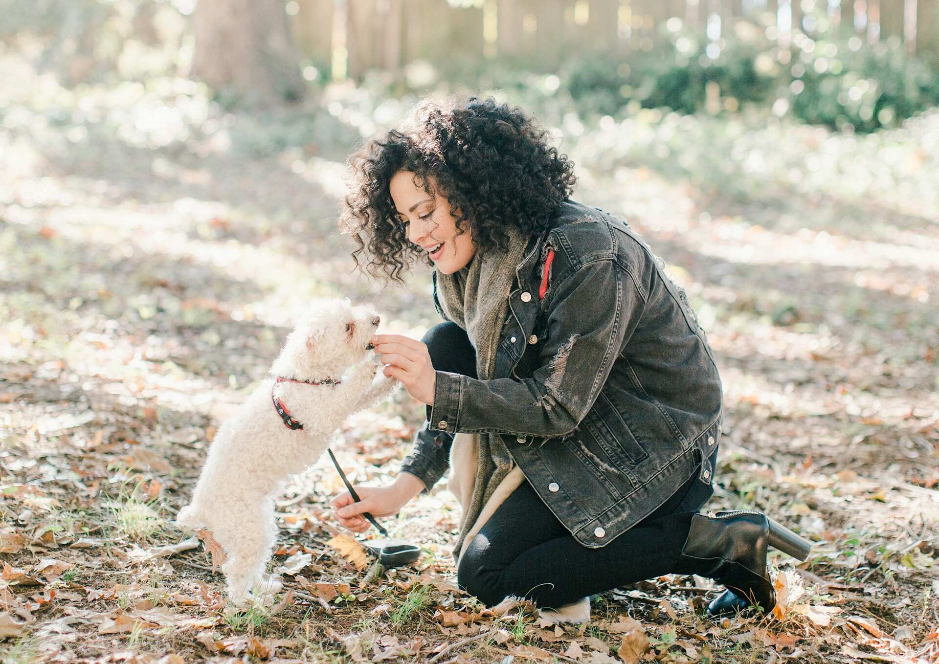 A woman feeding a small white dog a treat while out walking in a forest