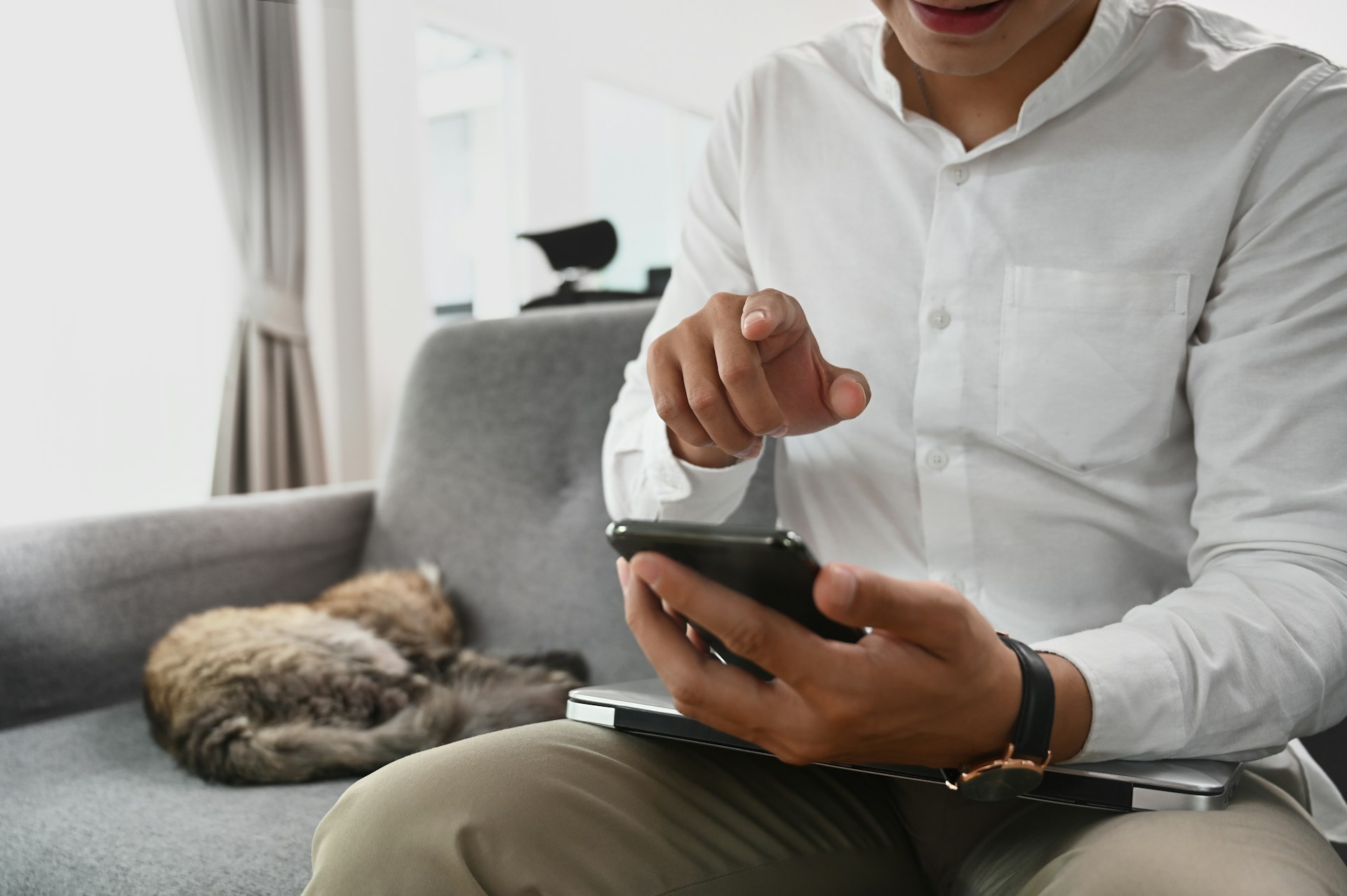 A man checking his phone sitting on a grey couch next to a cat